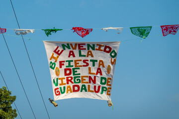 Oaxaca, Mexico. Dec 12, 2025. Large banner reading in Spanish "WELCOME TO THE FESTIVAL OF THE VIRGIN OF GUADALUPE" strung across the street on a clear, blue sky day.