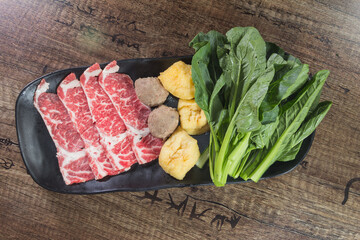 A hot pot platter with raw beef slices, meatballs, tofu puffs, and leafy greens arranged on a black plate.