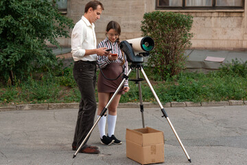 A young astronomy student stands beside her instructor while holding a telescope component, as the teacher explains its use during an outdoor scientific training session near observatory grounds