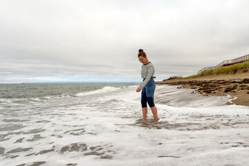 A young woman in striped top and rolled jeans enjoys walking barefoot in foamy ocean surf, blending human presence with natural coastal scenery on a breezy shoreline. Sea of Okhotsk Sakhalin Island