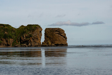 A closer view of a dramatic coastal rock formation split apart by erosion, with steep cliffs rising above the calm sea under a lightly clouded sky. Tikhaya Bay in the Sea of Okhotsk, Sakhalin Island