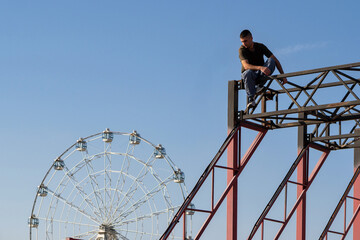 A male freerunner rests on a high beam overlooking a Ferris wheel in the background. The moment captures a mix of contemplation, daring, and visual contrast in an urban park