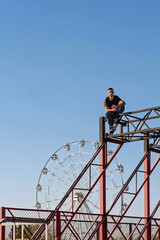 A male freerunner squats at the edge of a steel frame, framed visually by a Ferris wheel in the background. The composition highlights balance, geometry, and urban athleticism