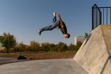 Male freerunner spins midair performing a backflip from a concrete ramp during parkour training in an outdoor park, under a bright blue sky, highlighting dynamic motion and agility