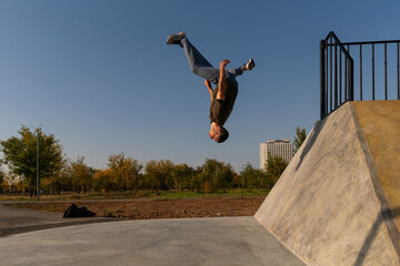 Male freerunner captured upside down mid-backflip off a concrete wall during parkour training in a sunny outdoor park, emphasizing body control, motion, and athletic discipline