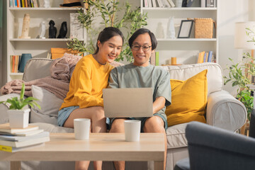 Asian married couple spends quality time together on a sofa in their living room at home, cheerfully using a laptop and smartphone, reflecting happiness and modern digital connection.