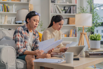 Asian married couple sits on their sofa at home, stressfully calculating financial budget and money loan accounts, reflecting unhappiness and debt pressure