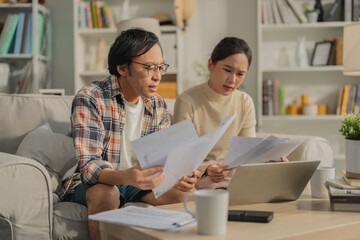 Asian married couple sits on their sofa at home, stressfully calculating financial budget and money loan accounts, reflecting unhappiness and debt pressure