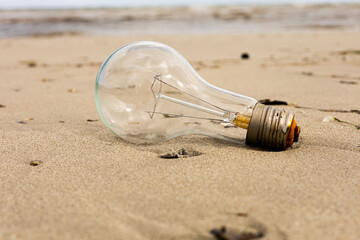 Obraz premium Big glass light bulb lying on sandy beach near the sea. Pollution waste, environmental awareness