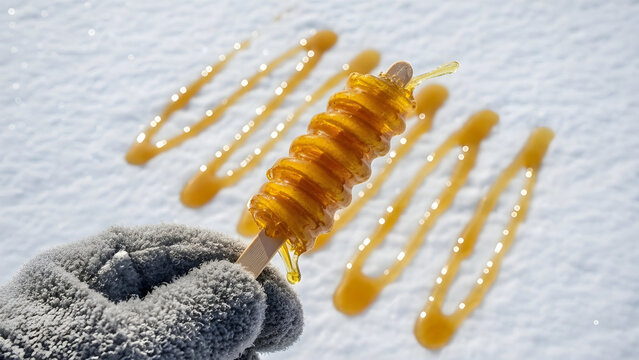 Gloved hand holding a dripping maple syrup taffy on a stick in snow
