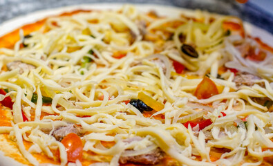 Close-up of an unbaked pizza topped with shredded cheese, tuna, onion slices, bell peppers and olives. Fresh ingredients and food preparation concept for cooking and restaurant use