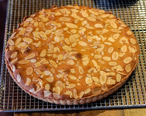 Golden brown almond tart topped with sliced almonds cooling on a metal rack. Homemade pastry, rustic baking, traditional dessert and bakery concept