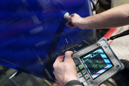 An aviation technician conducting ultrasonic testing on a blue aircraft rudder using portable NDT equipment with a digital display for defect detection, structural integrity inspection and maintenance