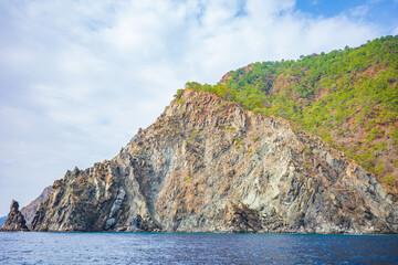 Striking coastal landscape featuring a rugged rocky cliff rising sharply from a calm deep blue sea, backed by lush green hills under a soft cloudy sky]