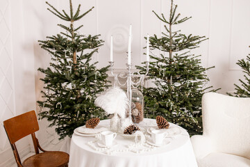 Elegant Minimalist Christmas Interior with White Table Setting, Pine Cones, and Festive Fir Trees Decorated with Lights and Bows in a Scandinavian Style