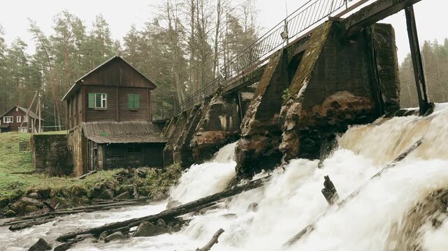 View of an old hydroelectric power station with a rushing river, historic dam, and a small wooden house nearby during overcast weather.