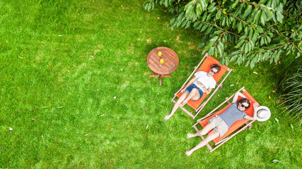 Young girls relax in summer garden in sunbed deckchairs on grass, women friends have fun outdoors in green park on weekend, aerial drone view from above
