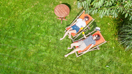 Young girls relax in summer garden in sunbed deckchairs on grass, women friends have fun outdoors in green park on weekend, aerial drone view from above