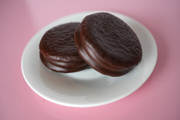 Two Chocolate-Coated Biscuits with Marshmallow Filling on a White Plate, Studio Shot on Pink Background.