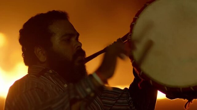 A slow-motion close-up of the hands and face of a drummer furiously playing the dhol drum, illuminated only by the dramatic light of a lohri fire against a dark background, a concept of the rhythmic e