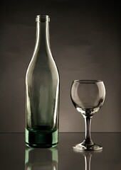 A clear, empty glass bottle stands next to an empty wine glass on a reflective surface against a dark background, showcasing their forms and transparency.