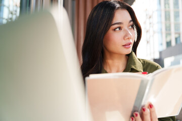 Young beautiful, elegant Asian woman in brown shirt hold notebook, looking away, thinking with computer laptop in luxury cafe. Digital technology, modern lifestyle, online learning and hybrid working
