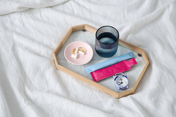 Tray with glass of water, vitamins and alarm clock on white bed.
