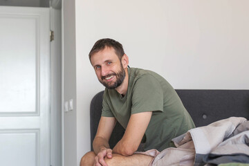 Fototapeta premium Adult man sitting on bed at home and smiling at camera, relaxed casual lifestyle in cozy bedroom interior, morning routine and everyday life concept, natural light and authentic domestic atmosphere