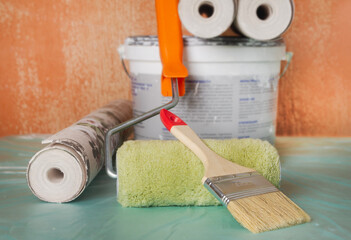 Wallpaper rolls, glue, paint, a roller, and a brush are ready for an apartment renovation. Room renovation tools are displayed against an old wall. Apartment renovation concept