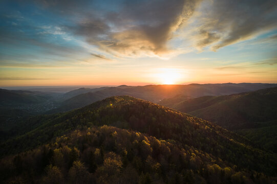Aerial view of breathtaking sunrise over rolling mountains covered in mix of lush green and autumnal foliage, with dramatic clouds streaking across sky, casting warm, golden light over the landscape.