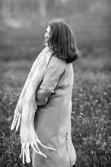 Young woman with long dark hair, wearing a stylish coat and scarf, stands confidently in a field, showcasing a serene expression and the beauty of nature in monochrome