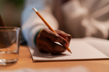 A person's dark-skinned hand holds a yellow pencil, writing in a grid-lined notebook. A glass of water sits nearby on the wooden table. This scene depicts focused work or study.
