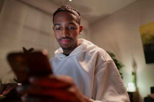 A young African American man in a white hoodie intently uses his smartphone in a warm, relaxed indoor setting. He is focused on his mobile device, browsing or communicating.
