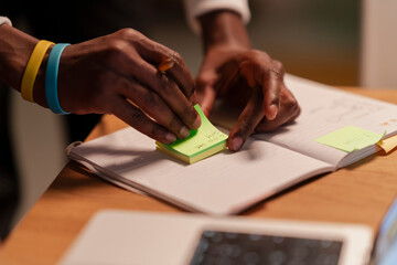 A person with colorful wristbands places a sticky note on an open notebook, symbolizing organization and planning.