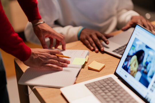 Two people collaborate at a desk, one writing in a notebook with a pencil, the other using a laptop, surrounded by sticky notes and another open laptop.