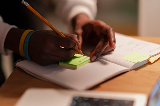 A person's hands write on a notepad with a pencil, with sticky notes and a tablet on the desk.