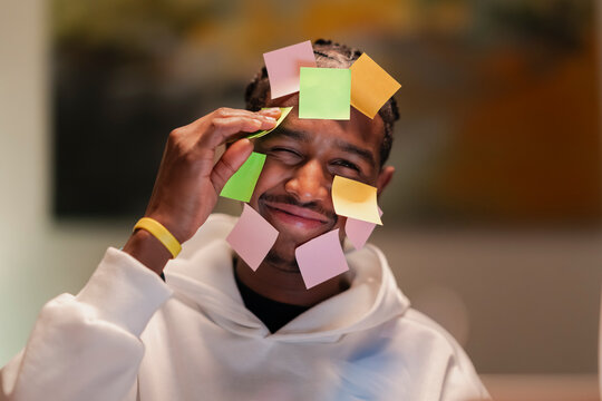 A young man with colorful sticky notes on his face and head smiles as he removes one from his forehead, conveying a sense of playful overwhelm or a creative brainstorming session.