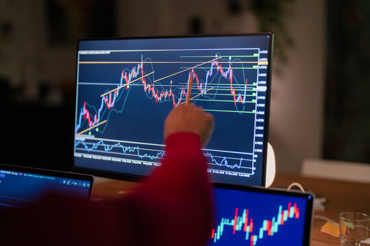 A person in a red shirt analyzes financial charts on multiple computer screens, using a pencil to point at the data.