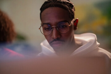 A young man wearing glasses and a hoodie looks intently at a screen, engrossed in his work or studies.
