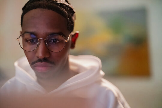 A young man with braided hair and glasses looks intently at a screen, his face illuminated by its glow.