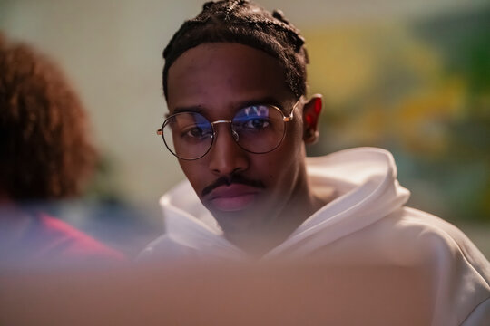 A young man wearing glasses and a white hoodie looks intently at a screen, his reflection visible in his lenses.