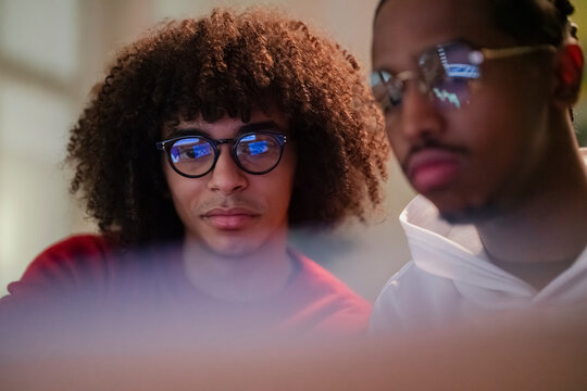 Two young men wearing glasses focus intently on a screen, their faces illuminated by its glow.