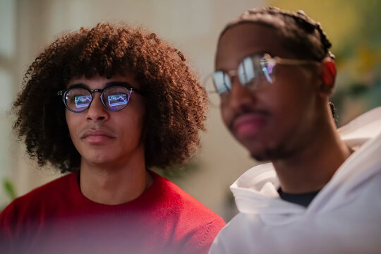 Two young men wearing glasses look intently at a screen, with a stock market graph reflected in one of their lenses.