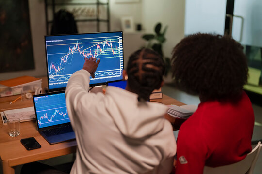 Two people analyze financial charts on computer screens, one person points to a detail on the main monitor.