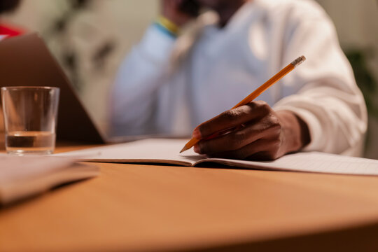 A person with dark skin writes in a notebook using a pencil on a wooden desk. A glass of water and a laptop are visible, suggesting a focused work or study environment.