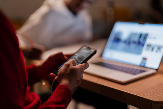 A person in a red sweater uses a smartphone while a laptop is open on a wooden desk, suggesting multitasking or studying.