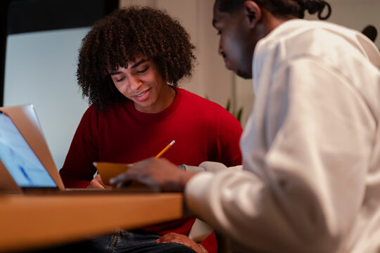 Two young men collaborate on a project, one using a laptop while the other takes notes with a pencil.