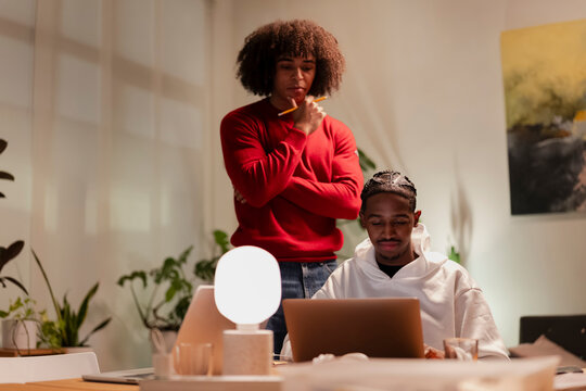 Two young men collaborate on a project, one focused on a laptop while the other observes thoughtfully, holding a pencil.