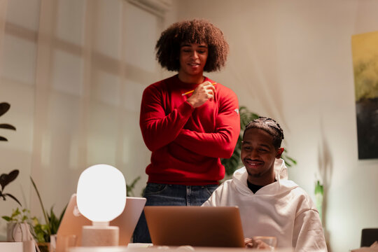 Two young men collaborate on a project, one looking at a laptop while the other stands nearby, holding a pencil and observing.
