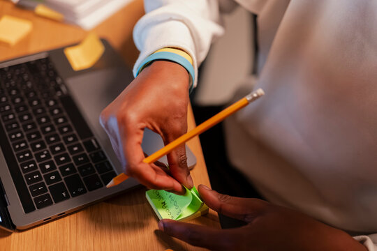 A person uses a pencil to write on a sticky note next to a laptop, organizing tasks and reminders for the day.